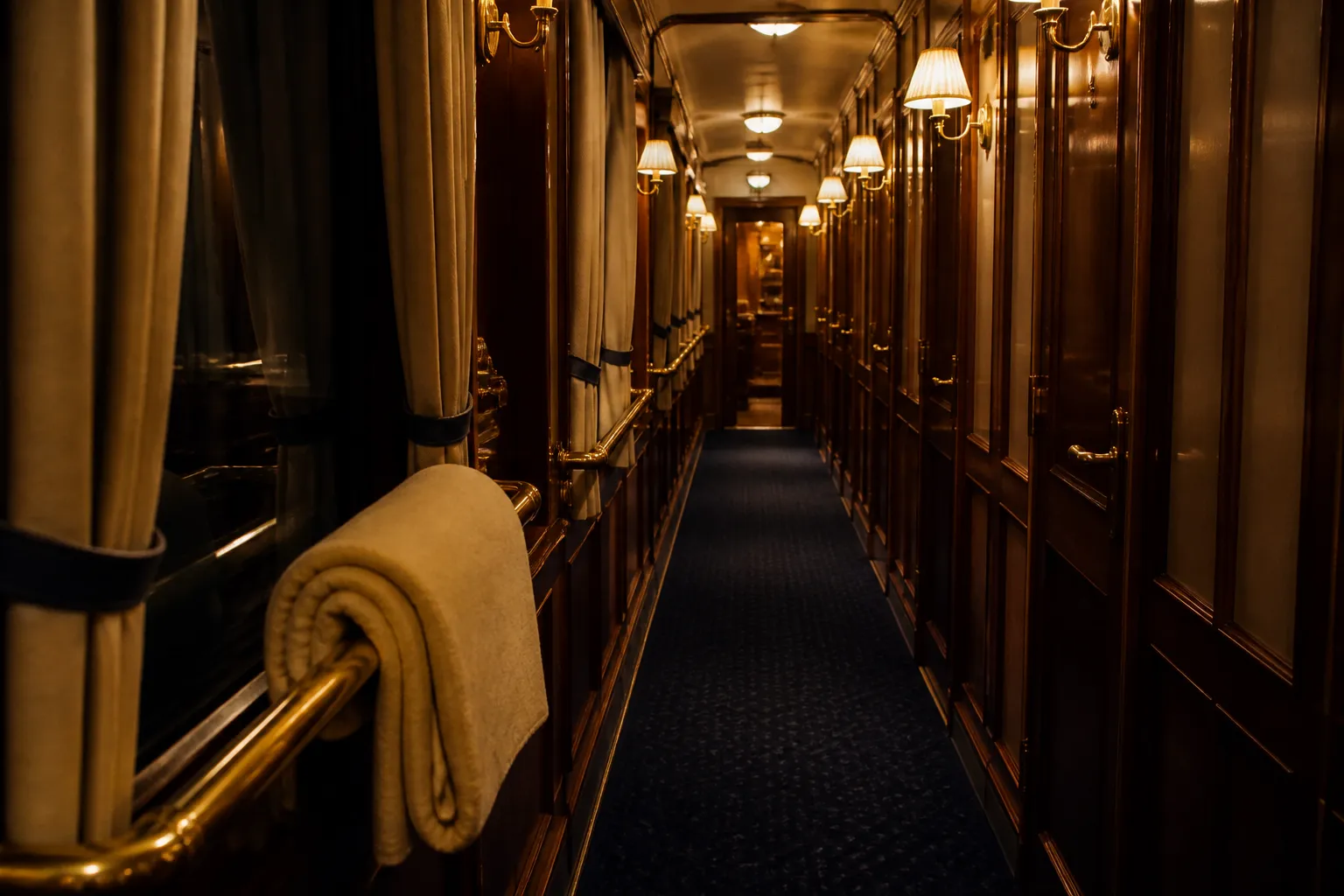 Narrow sleeper-car corridor at night, warm amber wall lamps lighting a row of closed compartment doors, dark blue carpet running toward a lit vestibule, a folded blanket resting on a brass handrail, 1920x1080